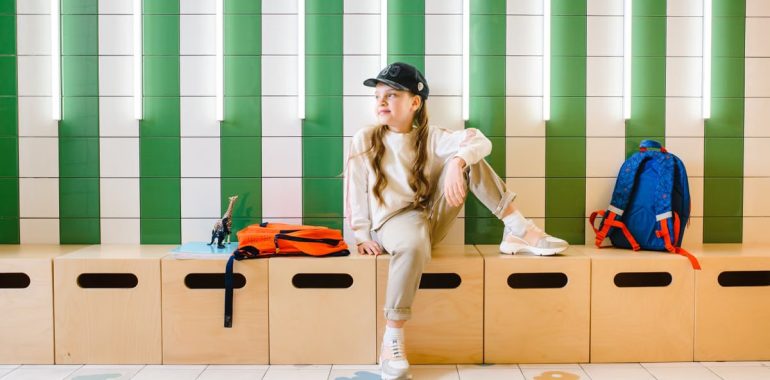 A young girl sitting indoors on a bench, with a backpack and hat, looking thoughtful.