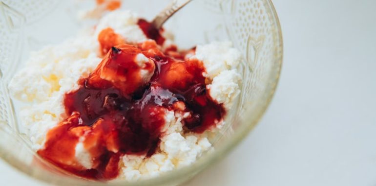 From above of appetizing cottage cheese with berry jam served in glass bowl with spoon on white table